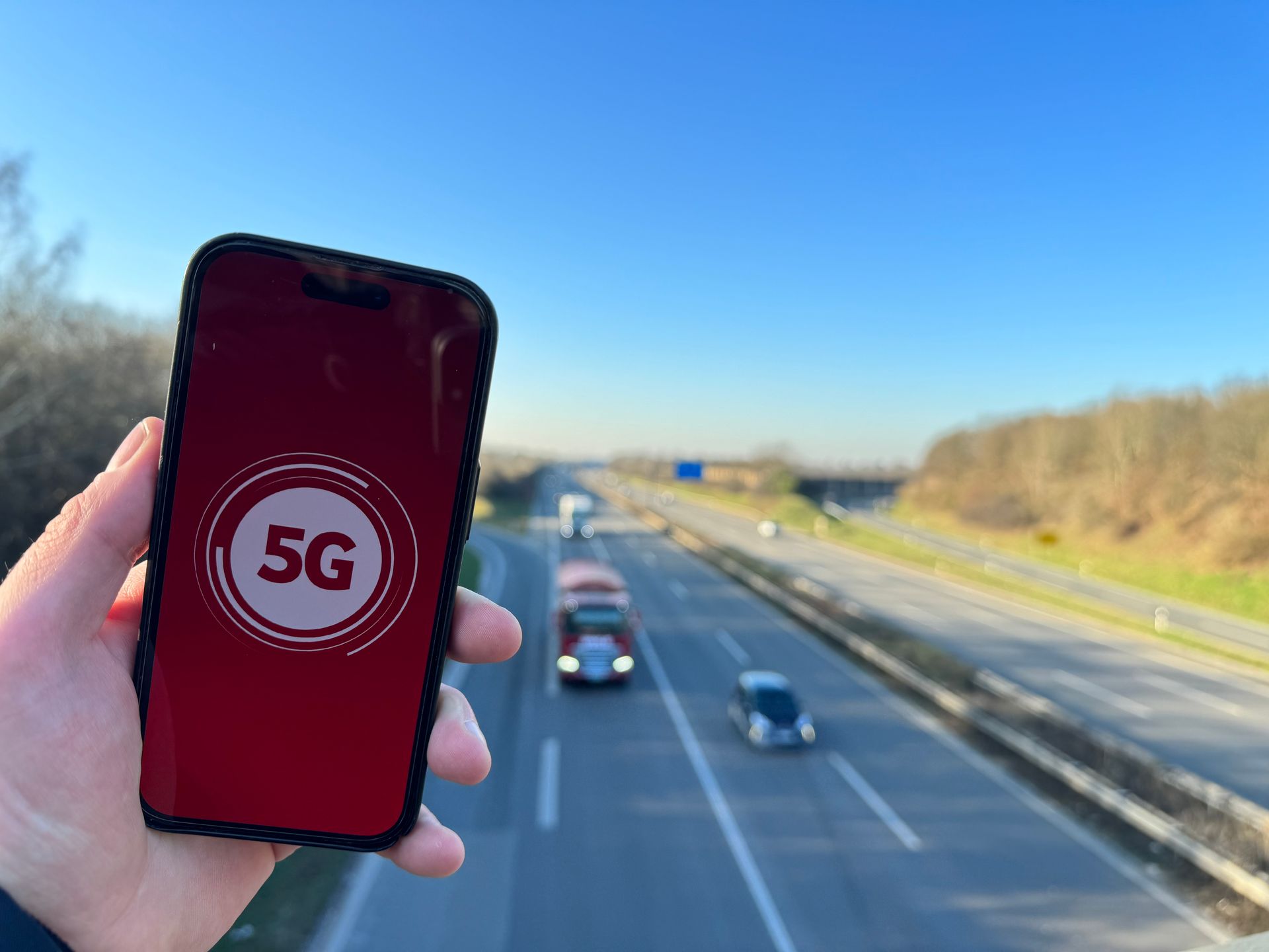 Blick von einer Autobahnbrücke, im Vordergrund eine Hand, die ein Handy mit 5G-Logo hochhält.