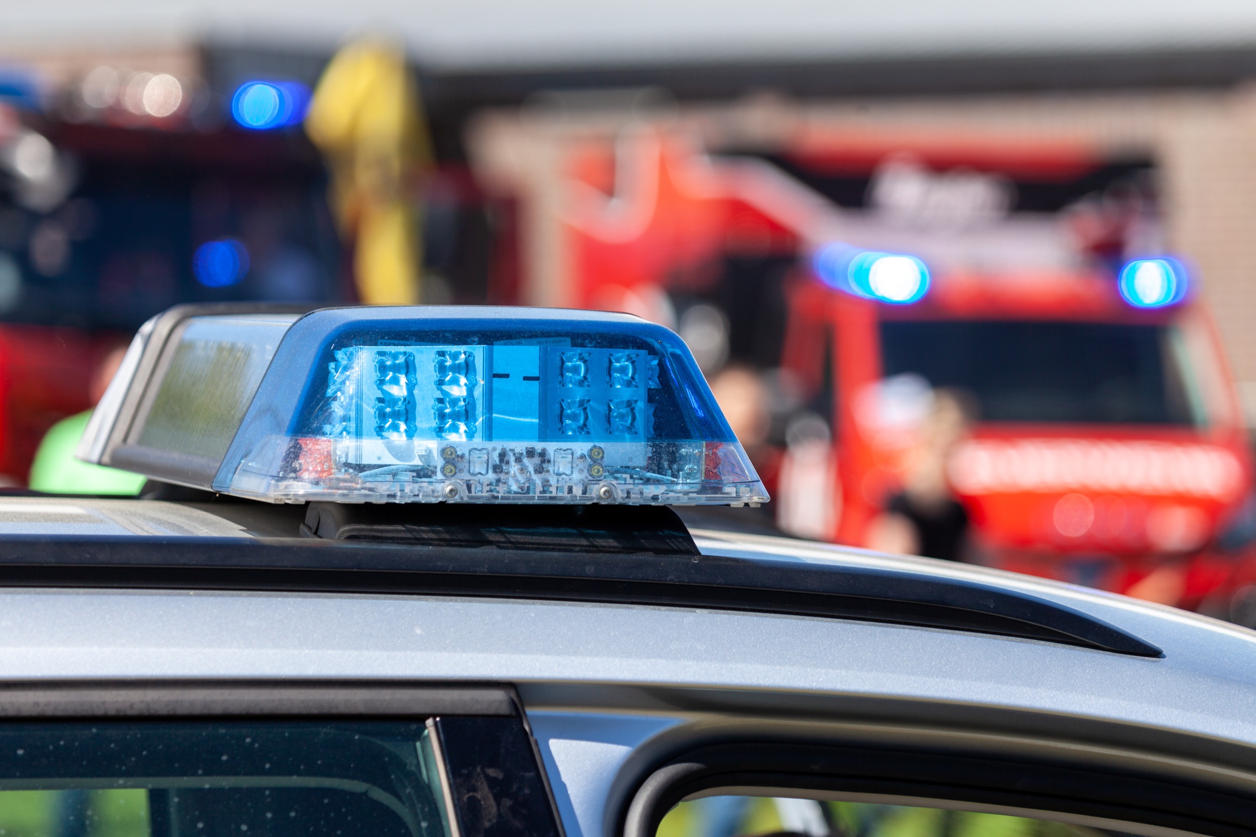Blaulichtbalken auf einem deutschen Polizeiauto, dahinter verschwommen Feuerwehrfahrzeuge mit eingeschaltetem Blaulicht.