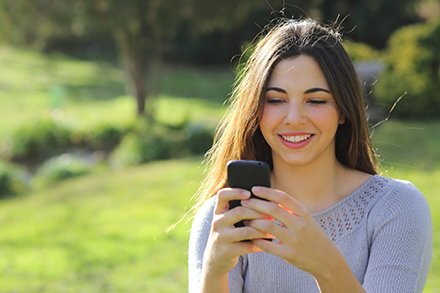 Eine langhaarige junge Frau steht auf einer Wiese und blickt lächelnd in ihr Smartphone.