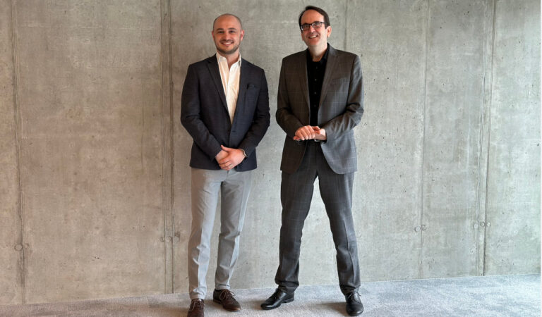 Zwei Männer in formaler Office-Kleidung stehen vor einer grauen Betonwand.