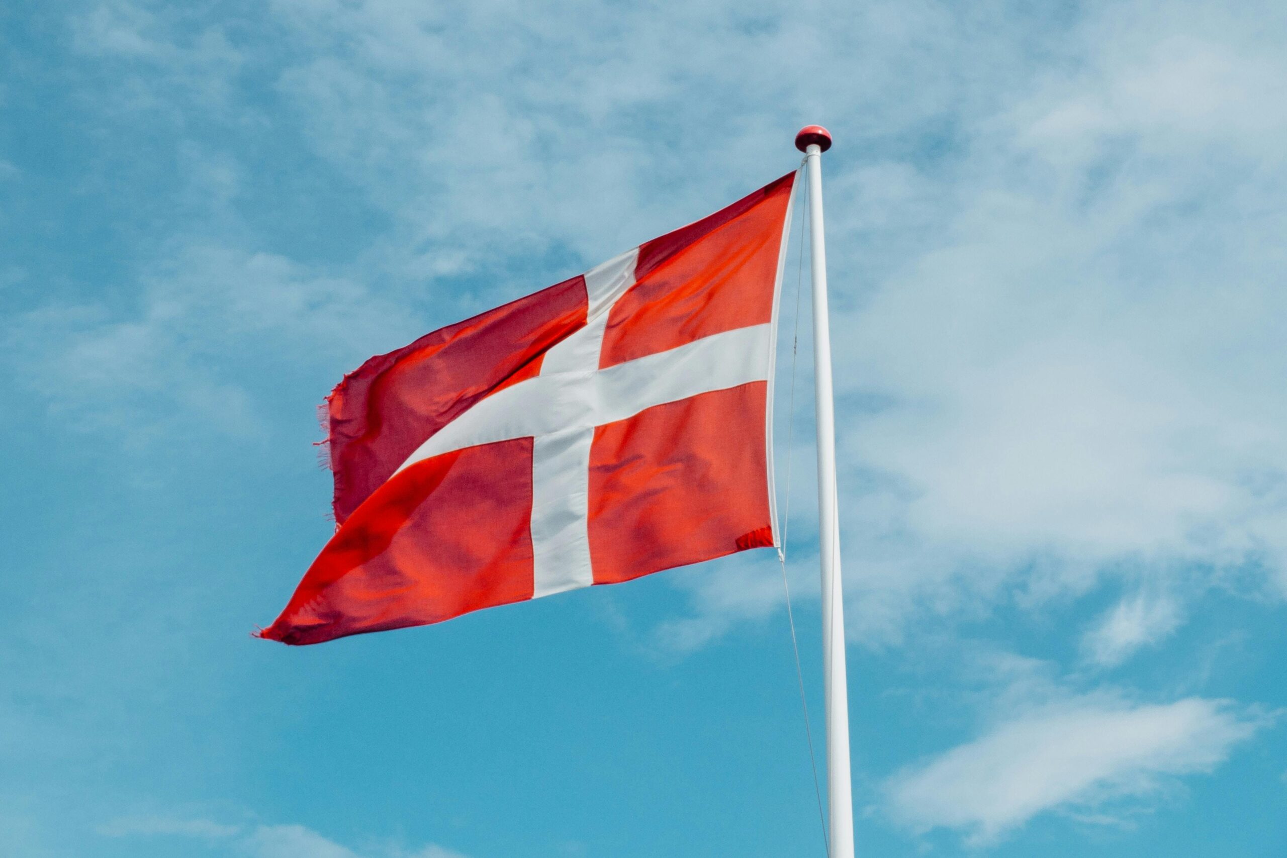Eine rote Flagge mit weißem Kreuz (Dänemark-Flagge) weht vor blauem Himmel von rechts nach links.