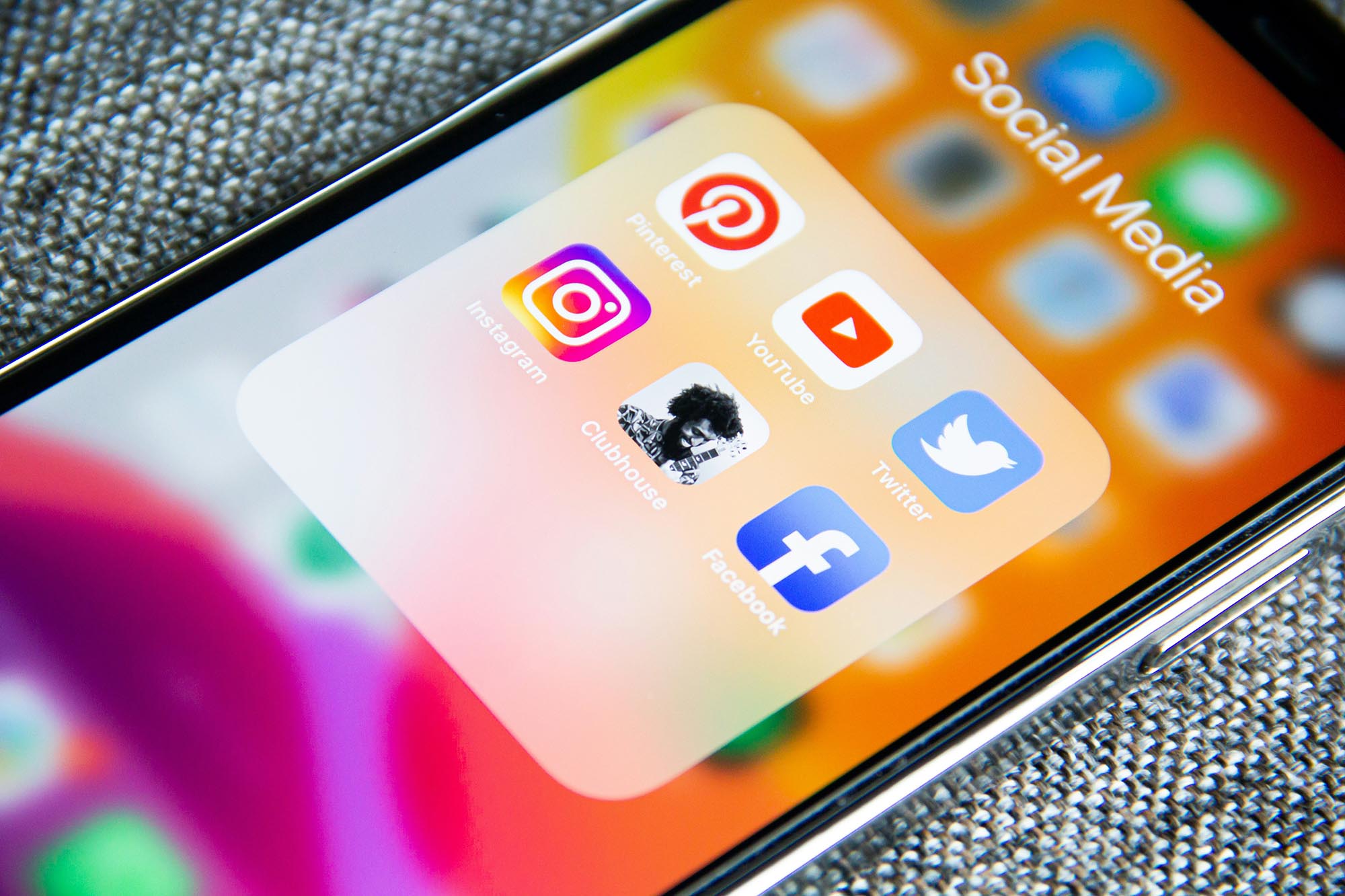 Eingeschaltetes Smartphone liegt diagonal auf einem grauen Stoff. Auf dem Screen sieht man Logos verschiedener Social-Media-Plattformen.