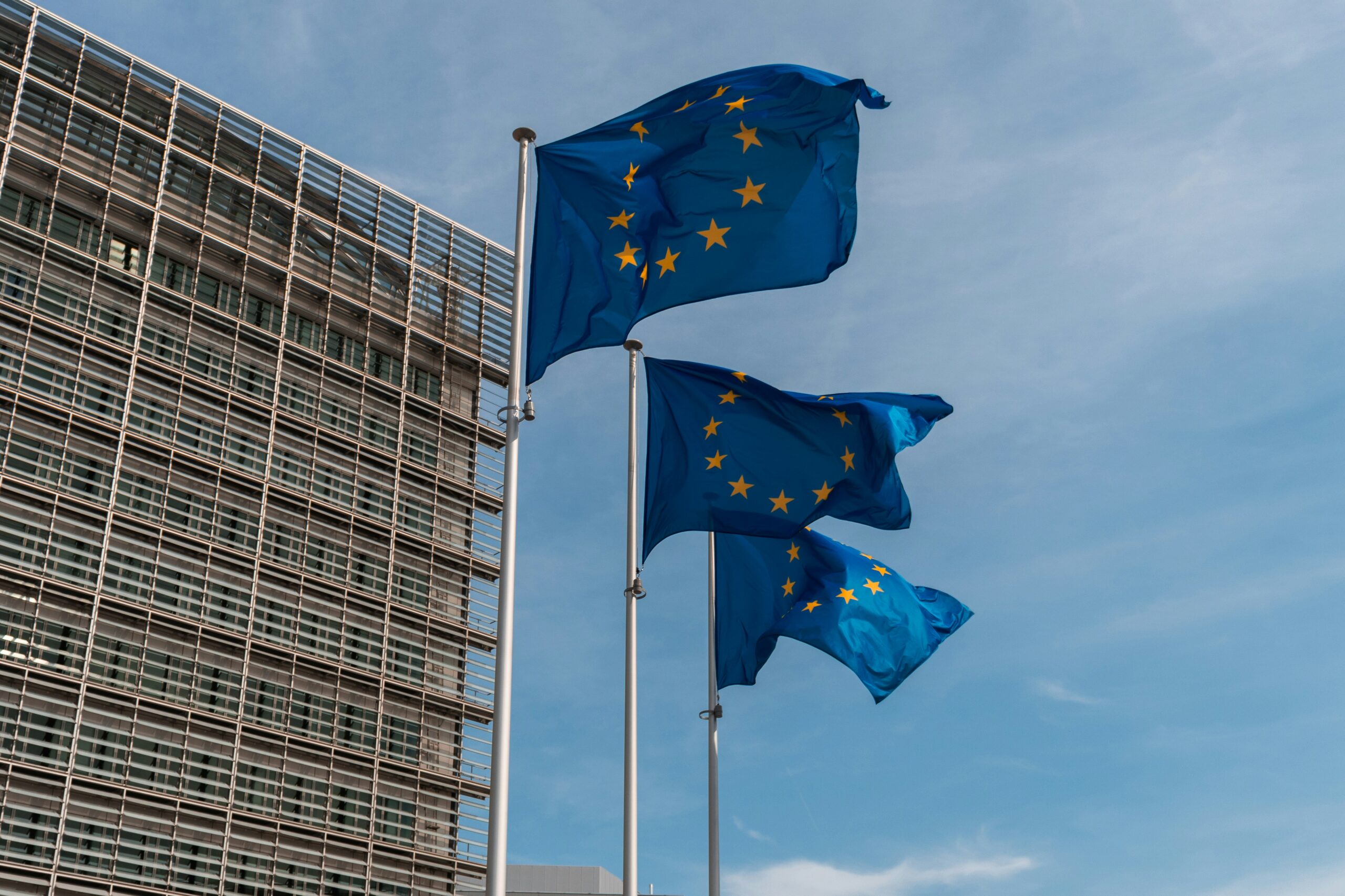 Drei EU-Flaggen vor blauem Himmel wehen im Wind, im Hintergrund ein modernes Gebäude und blauer Himmel.