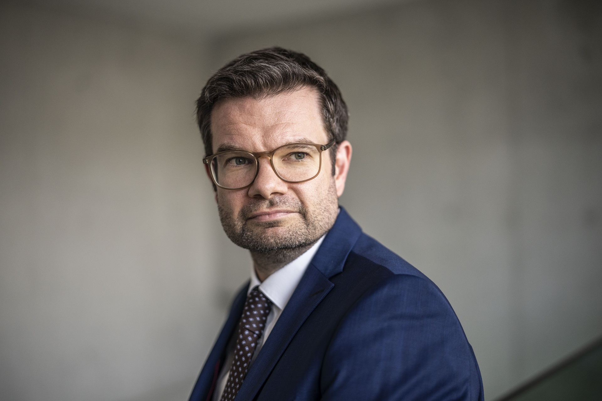 Porträtfoto von Bundesjustizminister Marco Buschmann im blauen Anzug, vor einer dunkelgrauen Wand.