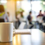 Eine Kaffeetasche steht im Vordergrund des Bildes auf einem Tisch, im Hintergrund sin schemenhaft Menschen zu erkennen, die zusammensitzen in einem großen Raum mit leeren Tischen.