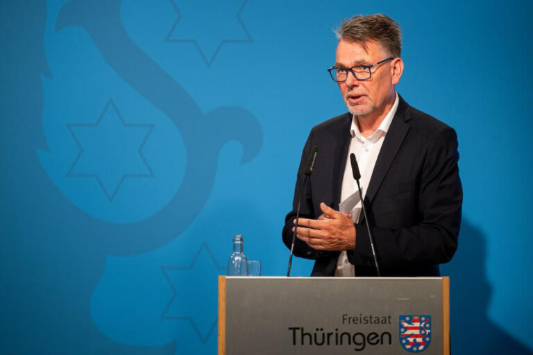 Thüringer Landes-CIO Hartmut Schubert im dunklen Anzug am Rednerpult, im Hintergrund eine blaue Wand.