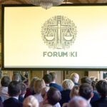 Blick über Köpfe und Schulter enes Publikums hinweg auf eine Vortragsbühne, dort ist das Logo "Forum KI" projiziert.