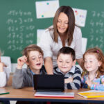 Gruppe von jungen Schulkindern sitzt vor einem Laptop, dahinter eine Lehrerin.