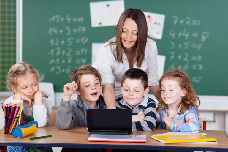 Gruppe von jungen Schulkindern sitzt vor einem Laptop, dahinter eine Lehrerin.