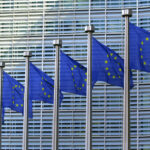 Eine Reihe von Flaggenmasten mit EU-Flaggen vor einer modernen Fassade.