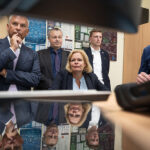 Blick unter einem Monitor hundurch über eine Tischplatte auf Bundesinnenministerin nancy Faeser, umstanden von mehreren Männern in Jackets.