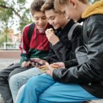 Drei männliche Teens sitzen draußen nebeneinander und schauen auf ein Handy, einer isst ein Butterbrot.