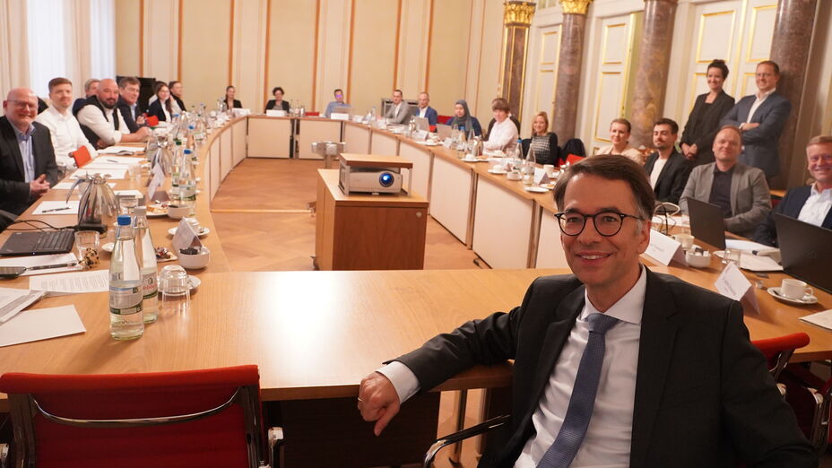 Blick in einen warm beleuchteten Raum mit Konferenztisch, an dem zahlreiche förmlich gekleidete Personen sitzen. Im Vordergrund ein Mann im Anzug, der freundlich in die Kamera sieht (Hessens Justizminister Christian Heinz).