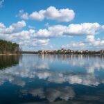 Blick über einen See, in dem sich der leicht bewölkte Himmel spiegelt. Am Ufer im Hintergrund ist eine Stadt zu erkennen.