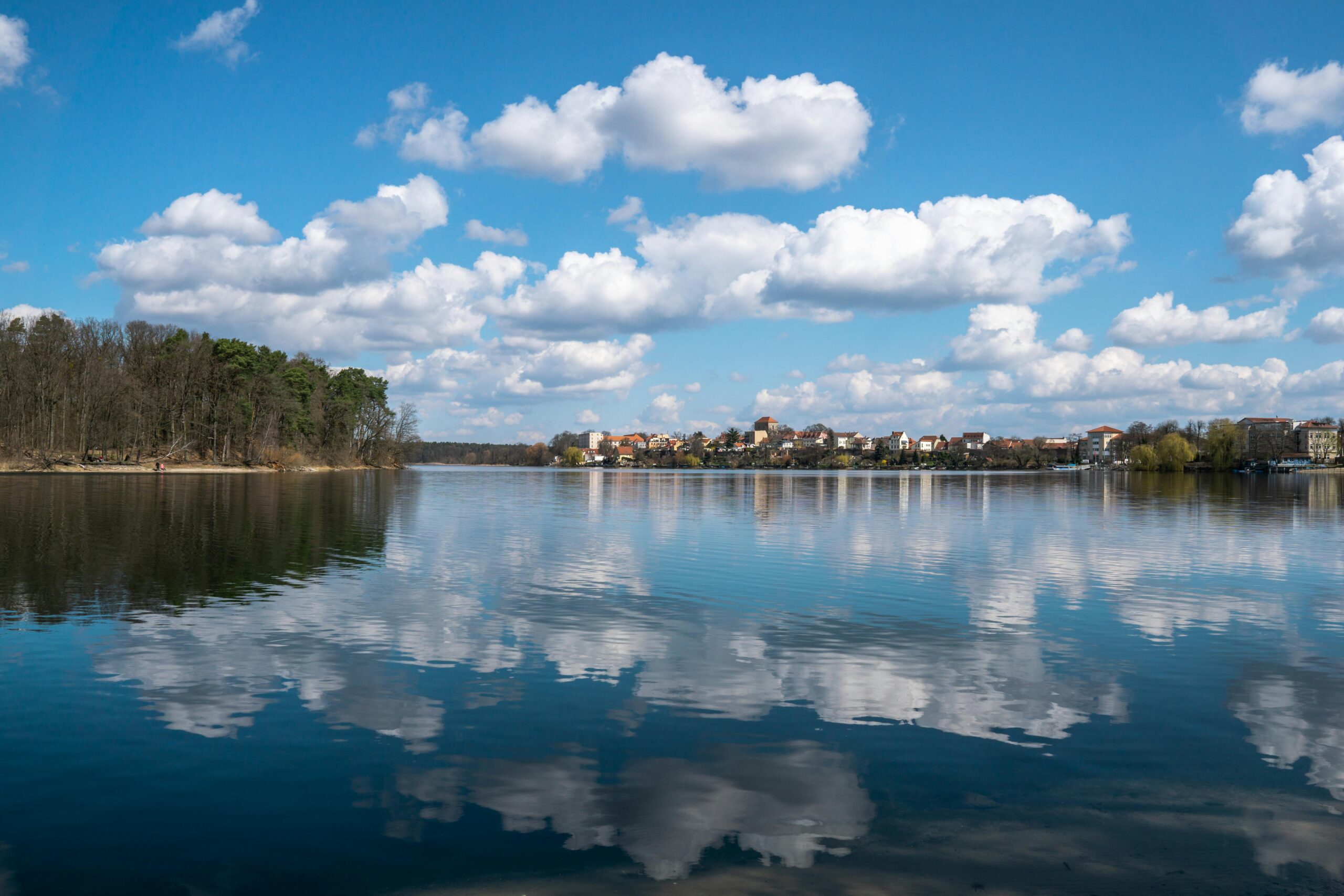 Blick über einen See, in dem sich der leicht bewölkte Himmel spiegelt. Am Ufer im Hintergrund ist eine Stadt zu erkennen.