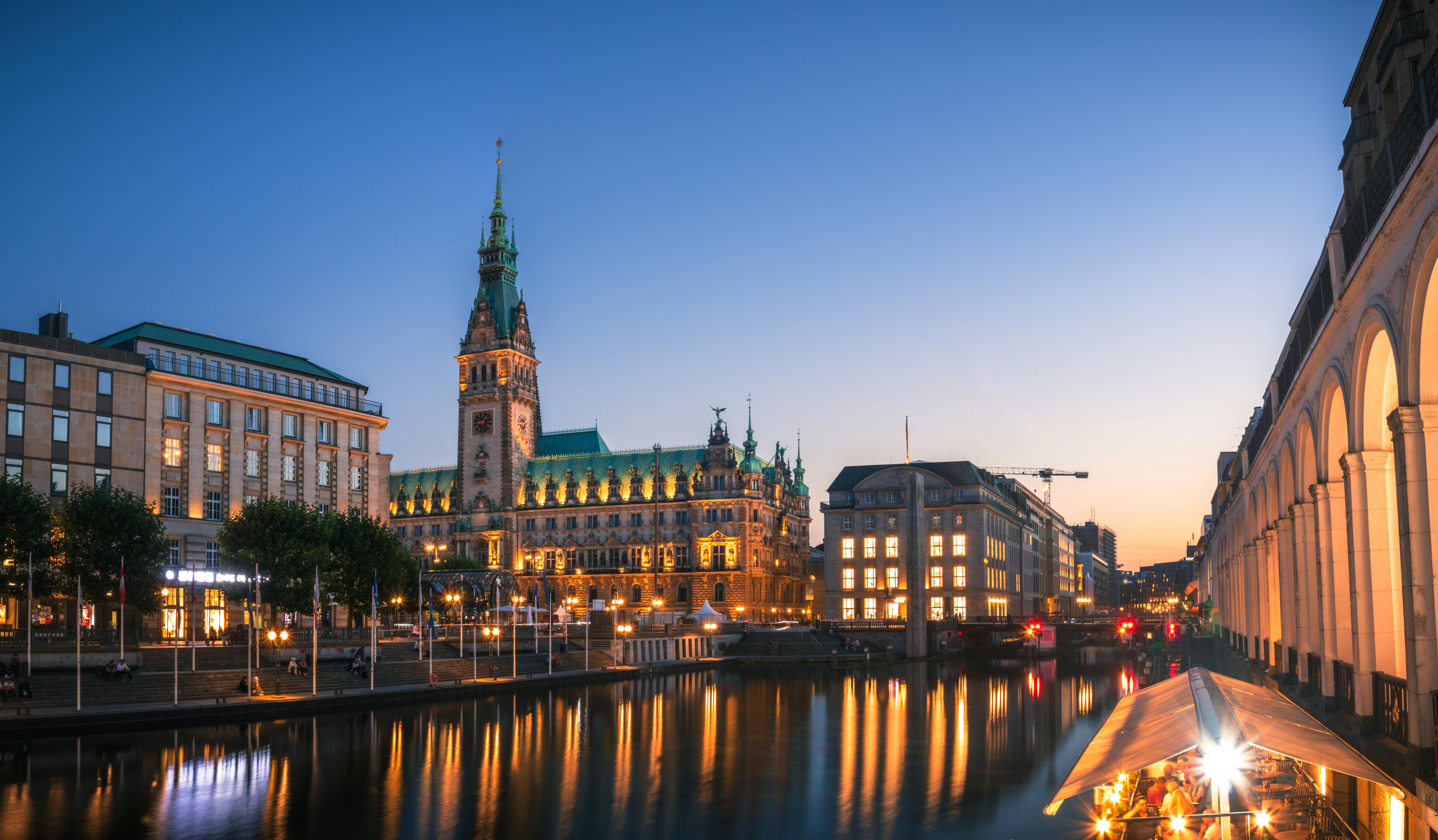 Ansicht der Innenstadt von Hamburg, im Vorergrund eine Wasserfläche, am Himmel Abenddämmerung.