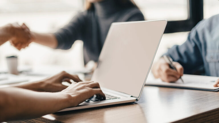 Nahaufnahme: Holztischplatte, ein paar männliche Hände arbeiten an einem Laptop, im Hintergrund sieht man unscharf und im Anschnitt weitere, eher junge Personen, eine schreibt etwas mit der Hand, es liegen Papiere herum.