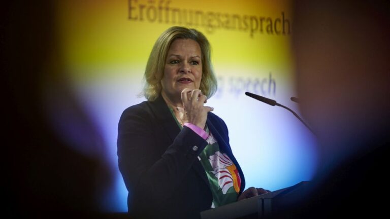 Bundesinnenministerin Nancy Faeser in dunkler, formaler Kleidung vor einem bunten Hintergrund, vor ihr ein schlankes Mikro.