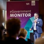 Ein bärtiger Mann im blauen Anzug mit Mikro auf einer Bühne, hinter ihm die Schrift "eGovernment Monitor".