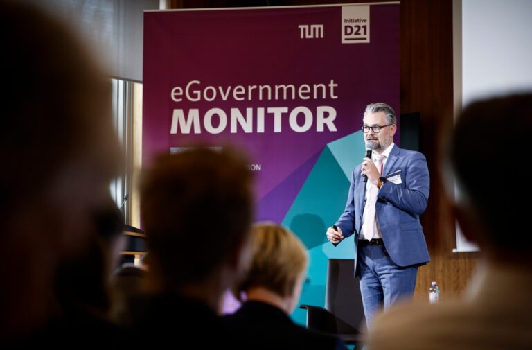 Ein bärtiger Mann im blauen Anzug mit Mikro auf einer Bühne, hinter ihm die Schrift "eGovernment Monitor".