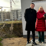 Außenafnahme einer Baustelle, am rechten Bildrand stehen ein Mann und drei Frauen und schauen in die Kamera.