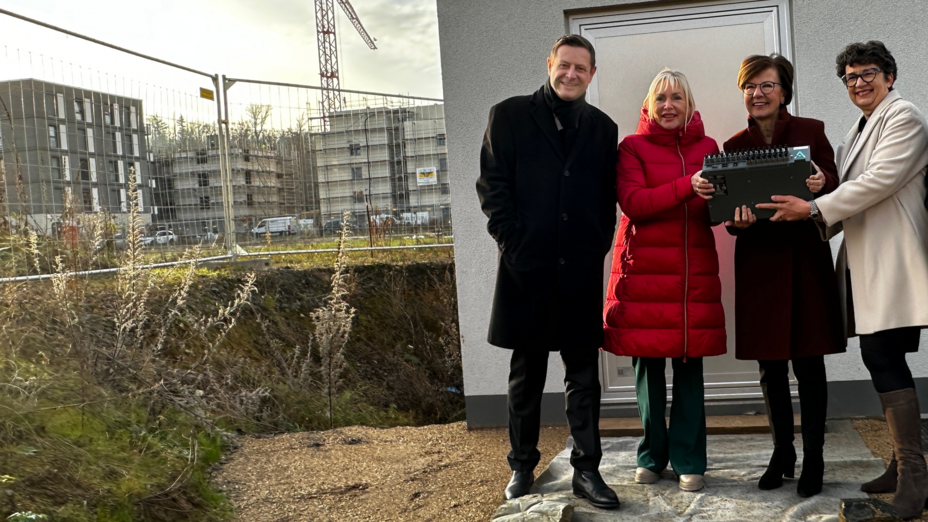 Außenafnahme einer Baustelle, am rechten Bildrand stehen ein Mann und drei Frauen und schauen in die Kamera.