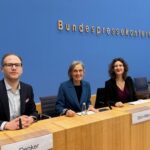 Ein Mann und zwei Frauen sitzen vor der blauen Wand der Bundespressekonferenz an einem langen Tisch mit Mikrofonen.