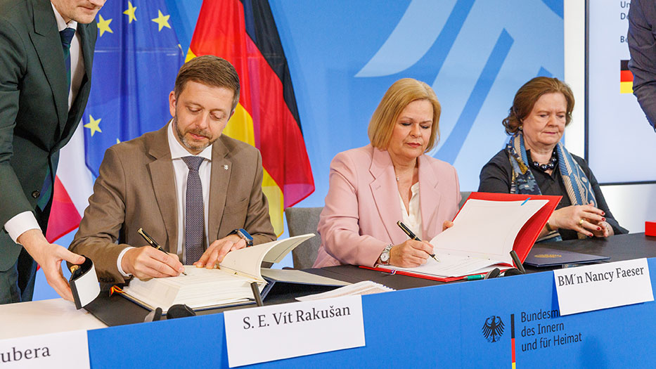 Vor dem Bundesadler und im Anschnitt sichtbaren EU-und Deutschlandfahnen unterzeichnen Rakušan und Faeser mit ernsten Gesichtern Papierdokumente.