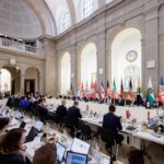 Regierungschefinnen und Regierungschefs der Länder mit Laptops an einem langen Konferenztisch in einem hohen, hellen Raum. Im Hintergrund klassizistische Kuppelarchitektur und die Landesfahnen.