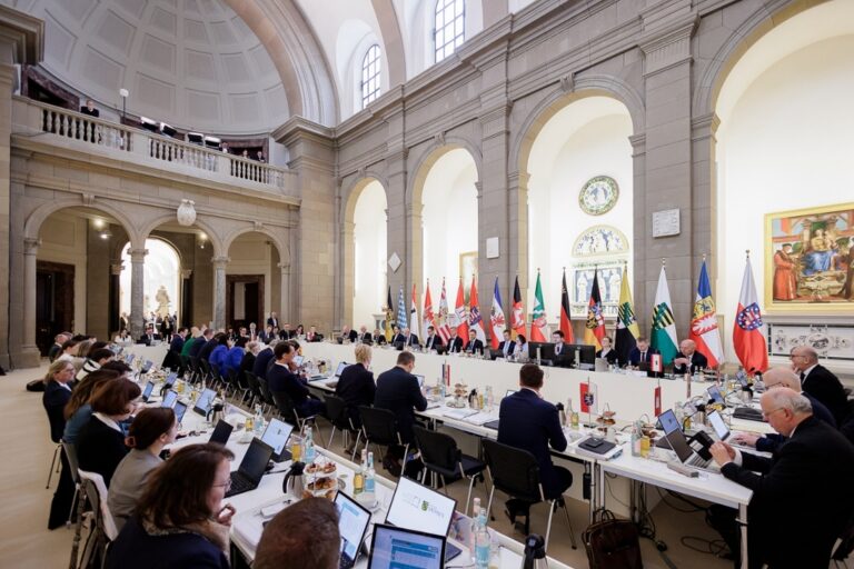 Regierungschefinnen und Regierungschefs der Länder mit Laptops an einem langen Konferenztisch in einem hohen, hellen Raum. Im Hintergrund klassizistische Kuppelarchitektur und die Landesfahnen.